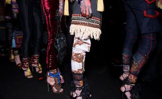 Close up shot of the lower half of several female models wearing looks from the Dsquared2 collection. One model is wearing black trousers. Another is wearing wine red coloured trousers with buttons. Next to her is a model wearing a blue, orange and off-white coloured piece with a fringe hem and multicoloured trousers with a zip. And the last model is wearing blue denim calf-length trousers with pockets and red detail. All models are wearing high heel sandals featuring tassels and lace