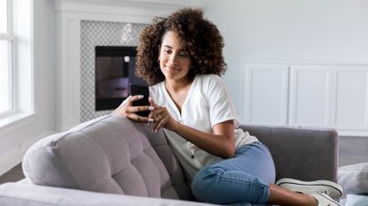 Woman smiles as she uses her phone while sat on a sofa.