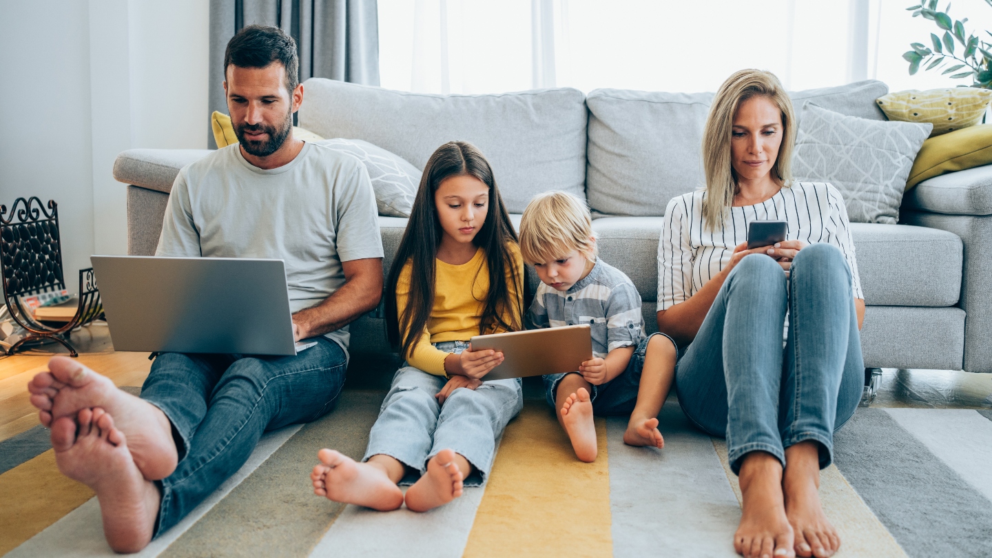 A family of four sitting on the floor in front of a couch all using technology