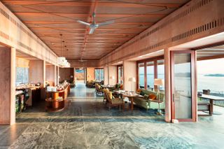 Daytime image of the RAAS Chhatrasagar restaurant, wooden slat ceiling, ceiling fans, grey marble floor, wooden frame entrance door and window frames along the side walls, dining tables and chairs, floor standing lamps, surrounding landscape view through the windows and open door
