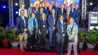 twenty-two well-dressed people wearing medals stand on a stage surrounded by potted plants