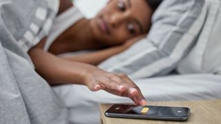 A woman in bed reaching over to her phone on a bedside table in order to turn off an alarm 