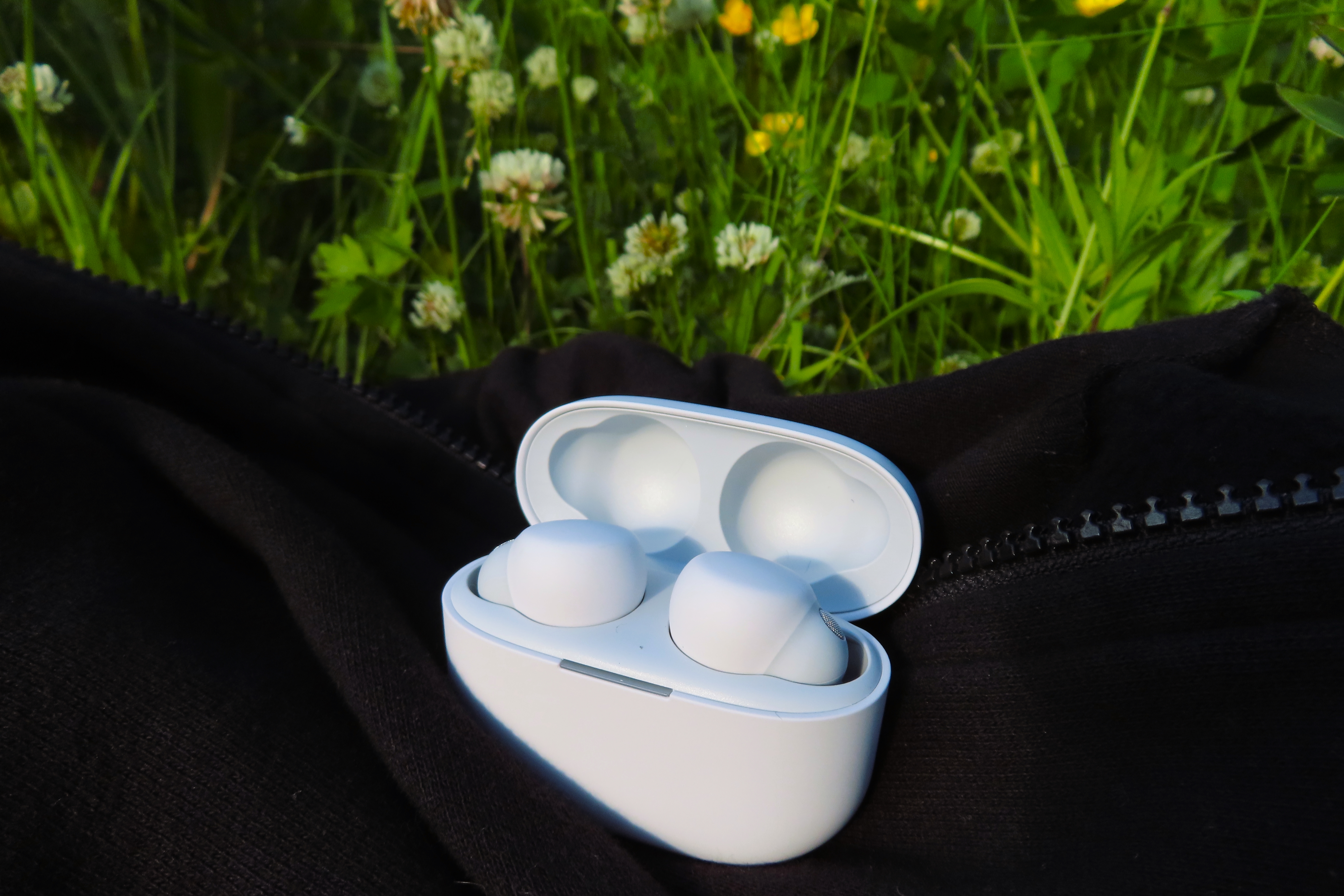 Ett par vita Sony LinkBuds S ligger i sitt laddningsfodral på en svart munktröja i högt gräs med blommor.