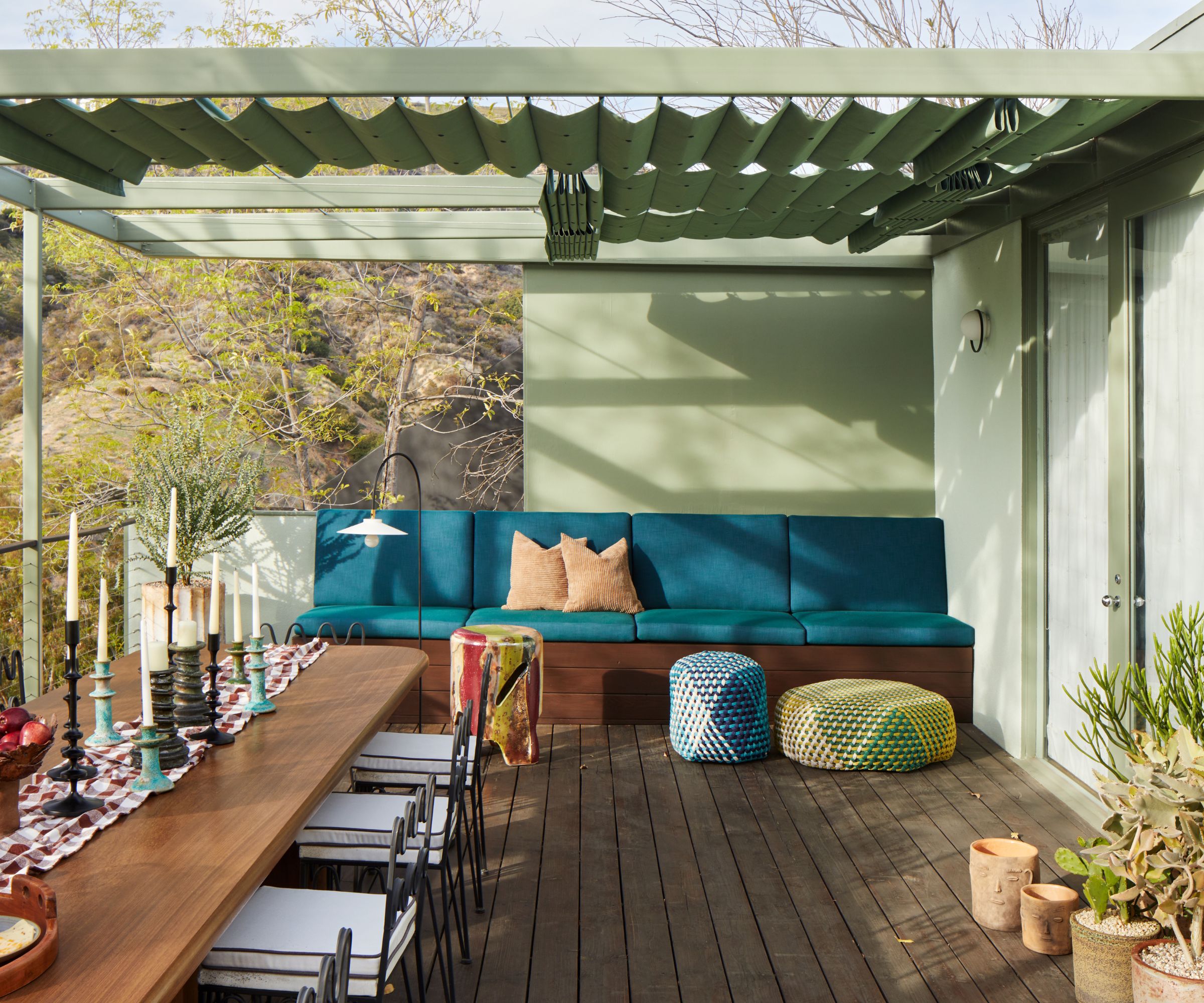 A balcony on a modern home with wood deck flooring, a sage green wall and roof, with a teal outdoor sofa, a large wooden dining table with dinner candles, and a table runner.