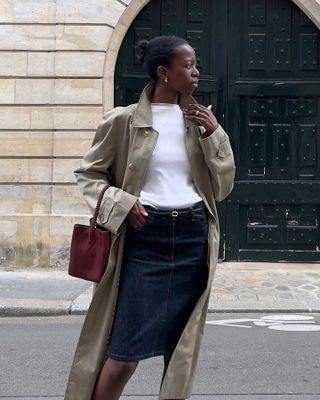 Paris based fashion influencer Sylvie Mus poses on the street in front of an ornate black door wearing a trench coat, white top, denim skirt, and burgundy bag