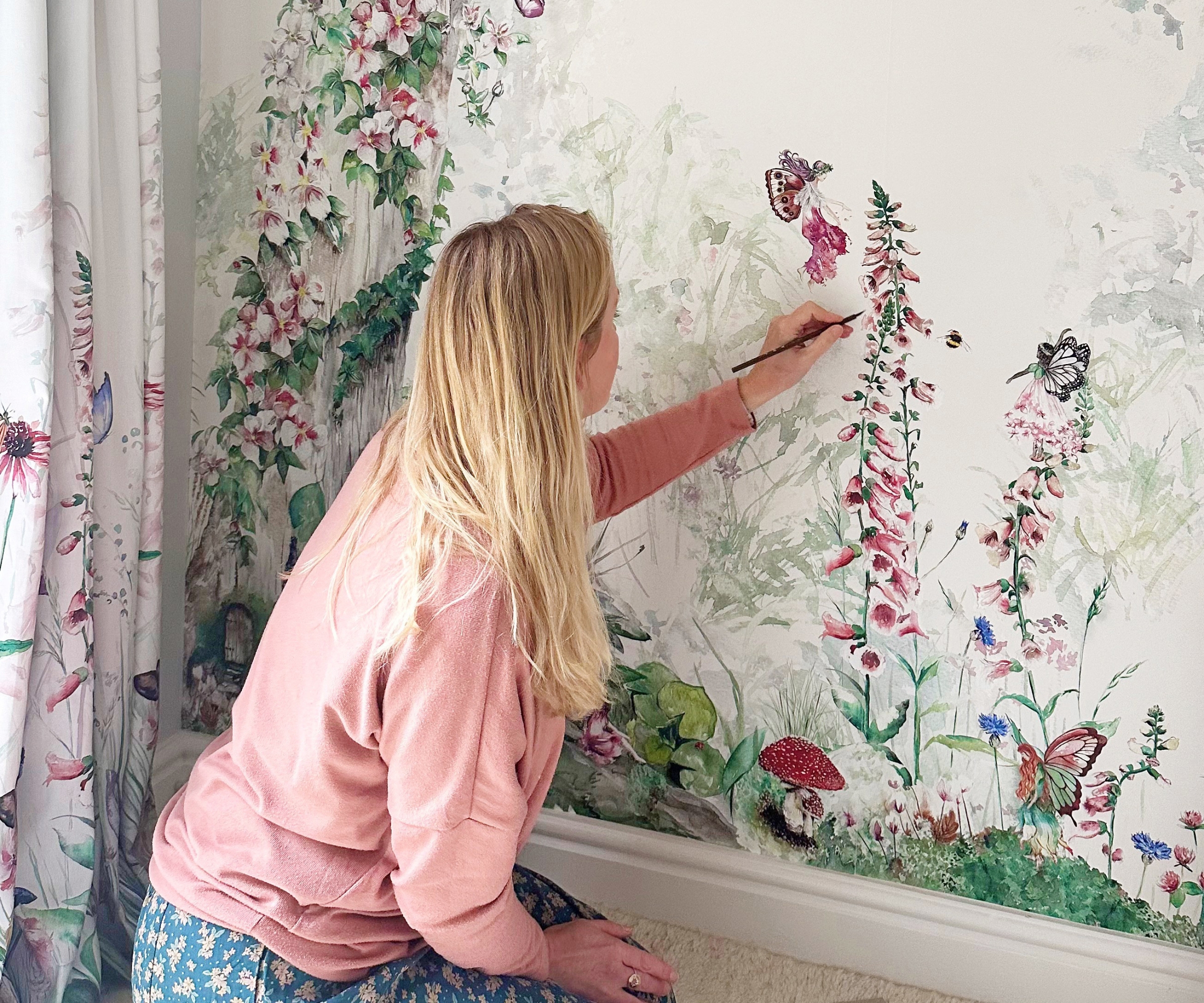 Woman with long blonde hair in pink jumper paints foxgloves on a floral fairy mural beside matching curtains