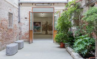 The entrace of the gallery with the double doors open. The walk way leading to the doors has two grey carved stone benches against a wall on the left and on the right green plants against a wall. Through the door is a peek of an abstract painting on the wall