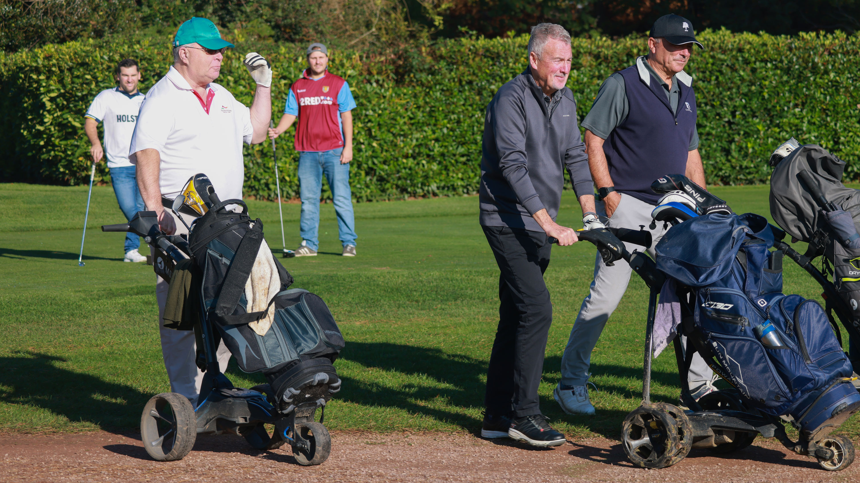 three traditionally dressed golfers walk off the tee while two other golfer in football kits and jeans wait behind