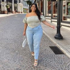 gia wears off shoulder green top and light wash straight cut blue jeans and white heeled sandals and carries a white purse