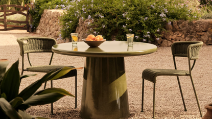 A round Reti Dining Table, Glazed, Olive with patio chairs on each side and a bowl of oranges with two glass cups 