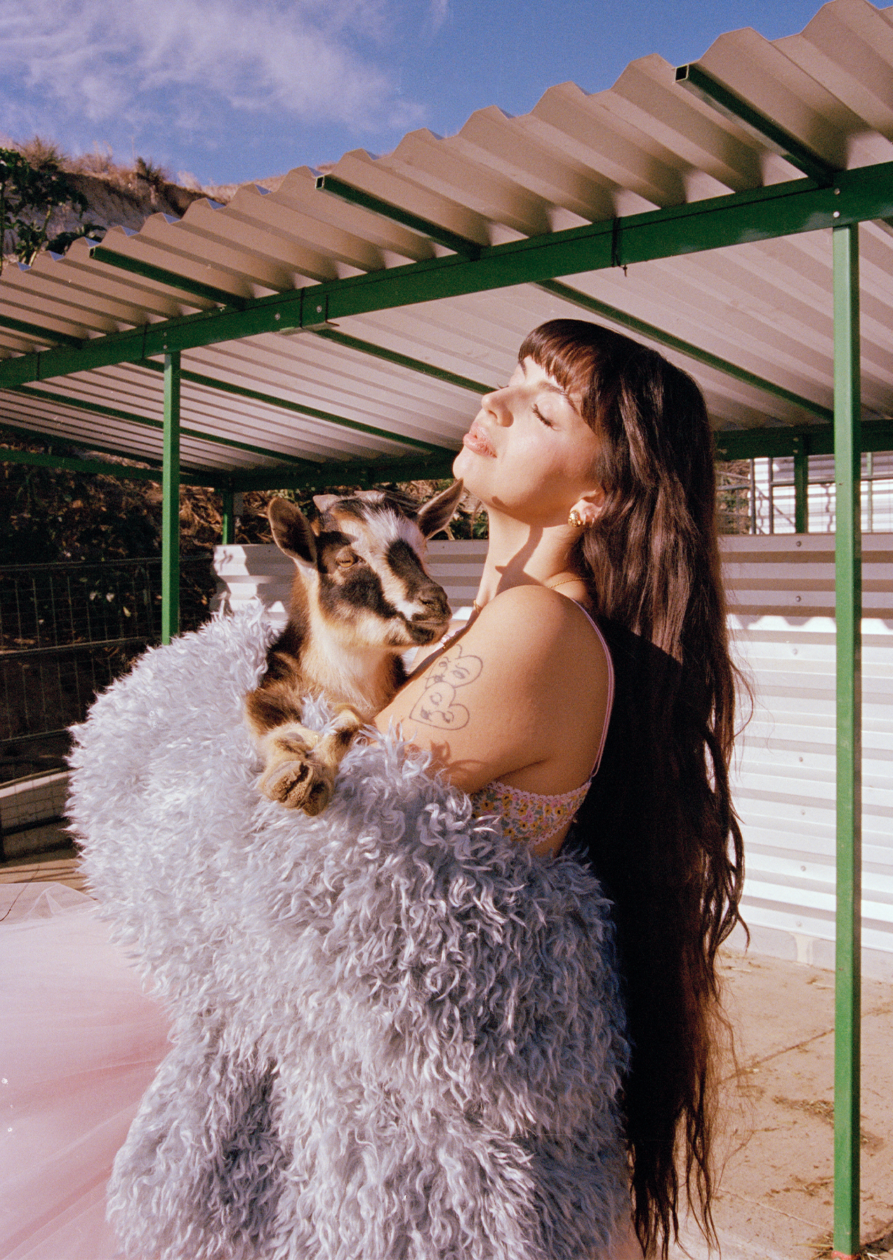 Singer Rebecca Black is on a farm holding a goat. Her bangs are short, her hair is long, and she has natural waves. She wore a pink flower bra with a furry gray coat.