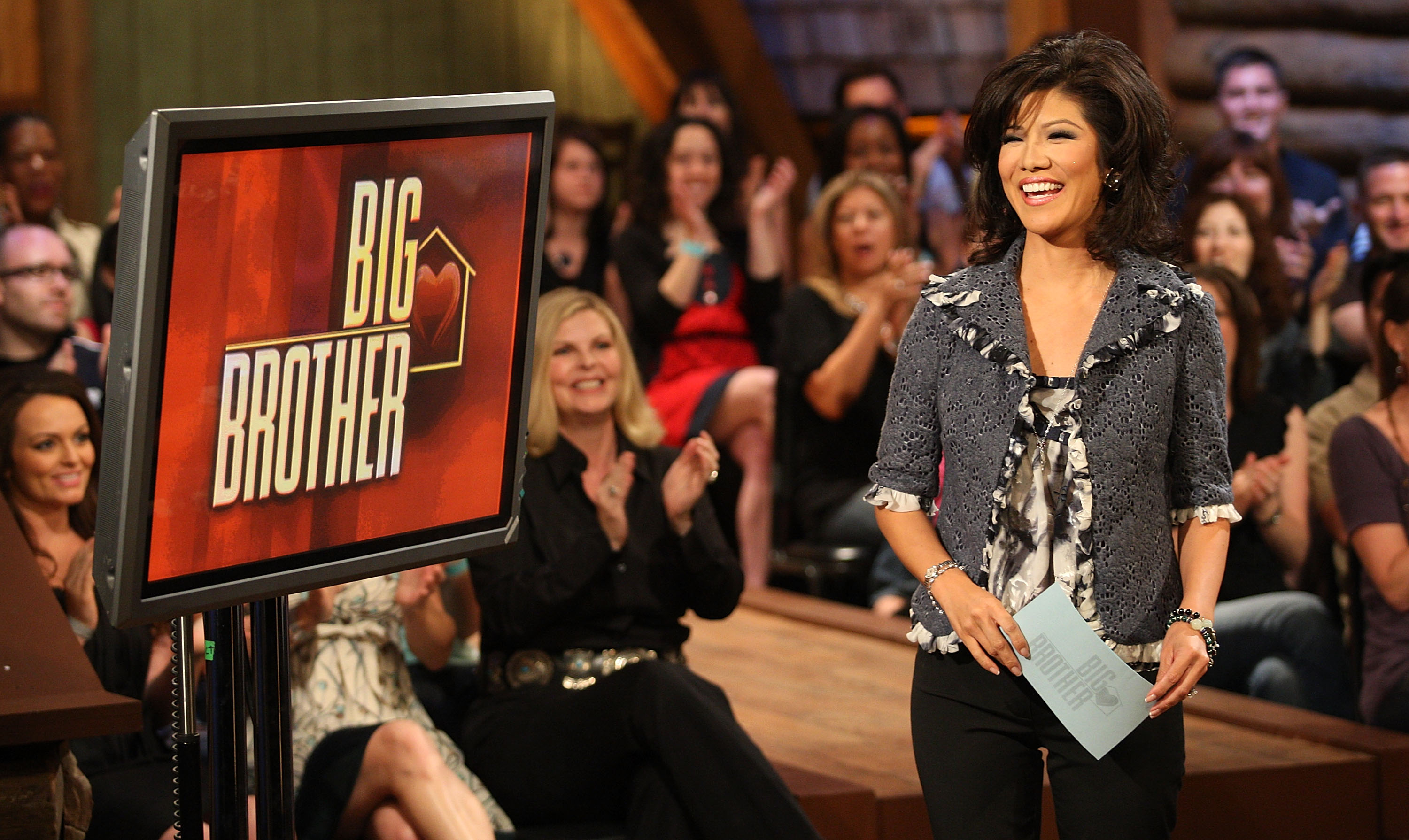 the host of big brother stands in front of an audience by a tv with the title on it