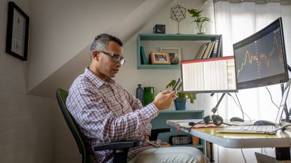 A man looks at a trading app on his smartphone while sitting at his desk in his home office.