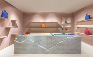 A store with handbags on the wall shelves and a grey brick display platform in the middle of the room with various sized and coloured leather wallets, purses and pockets on it.