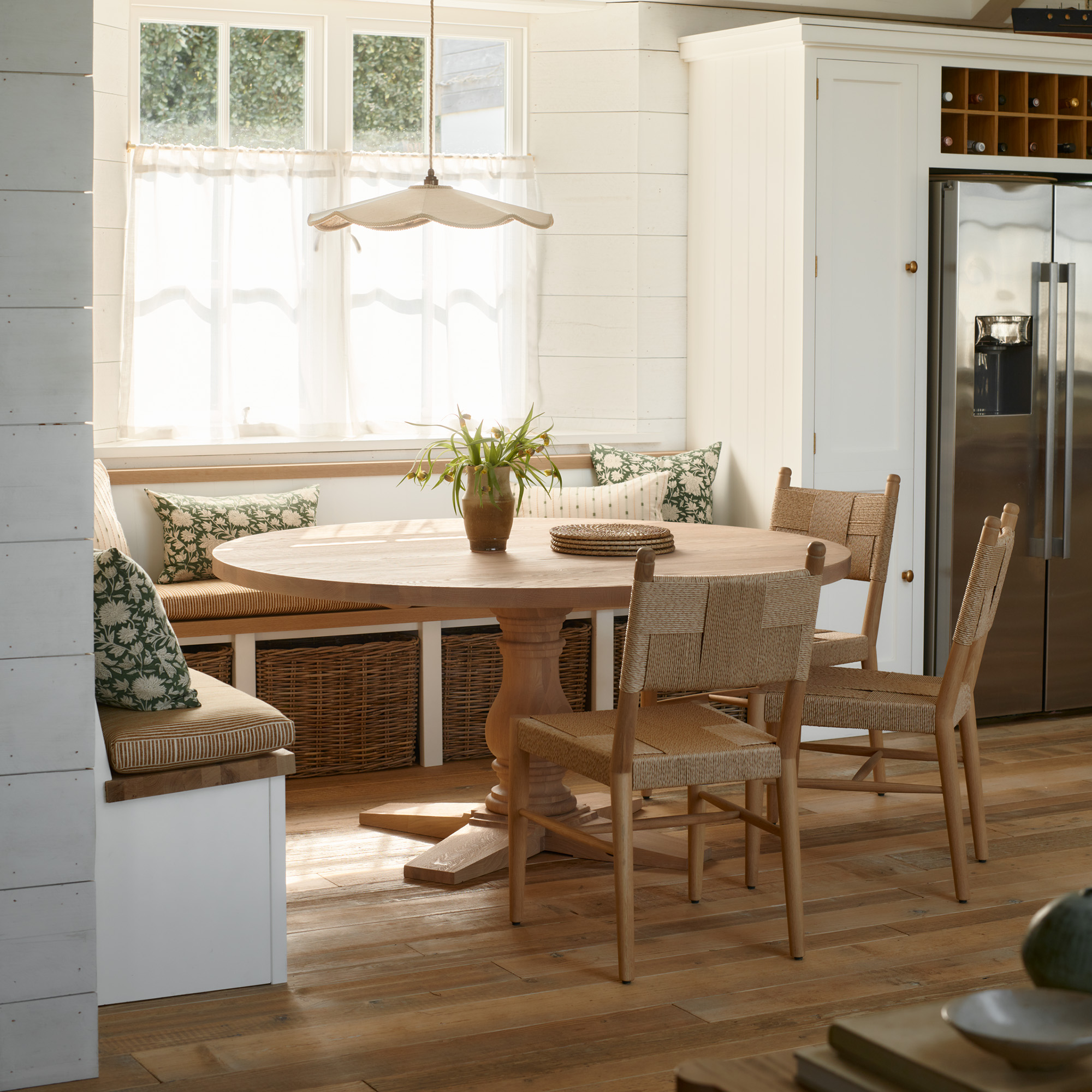 neutral dining room with round table, wooden chairs and banquette seating with rattan baskets a pendant light hangs above the table and the windows are partially dressed with a sheer linen curtain