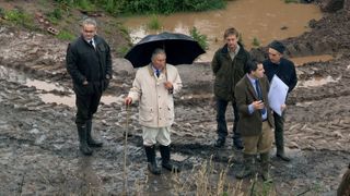 King Charles III in a mac and wellies and with an umbrella and stick walks through mud with his restoration team.