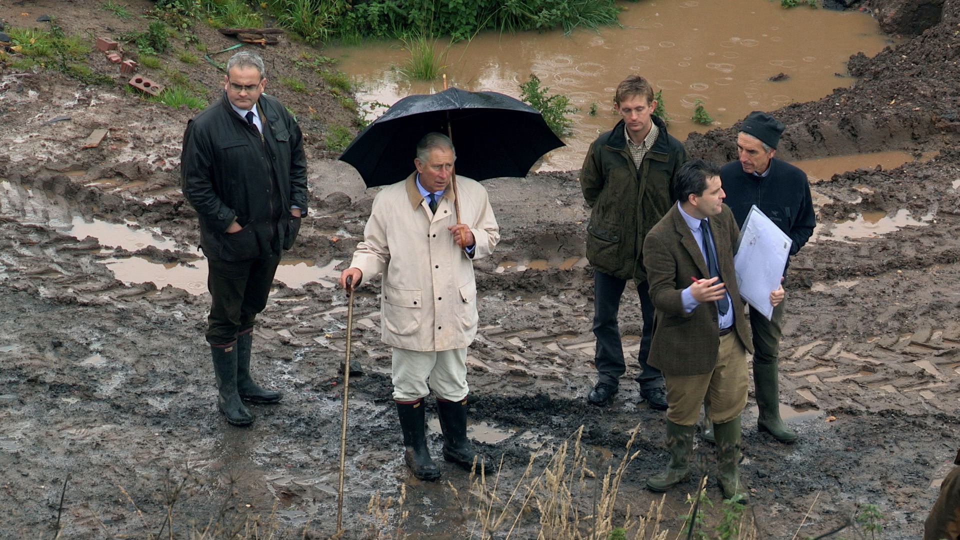 King Charles III in a mac and rubber boots and carrying an umbrella and cane walks through the mud with his restoration team.