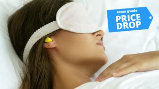 A woman with brown hair sleeping in bed with white sheets wearing a white sleep mask and yellow noise cancelling earplugs with Tom's Guide price drop logo