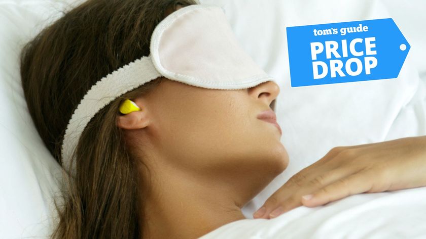 A woman with brown hair sleeping in bed with white sheets wearing a white sleep mask and yellow noise cancelling earplugs with Tom&#039;s Guide price drop logo