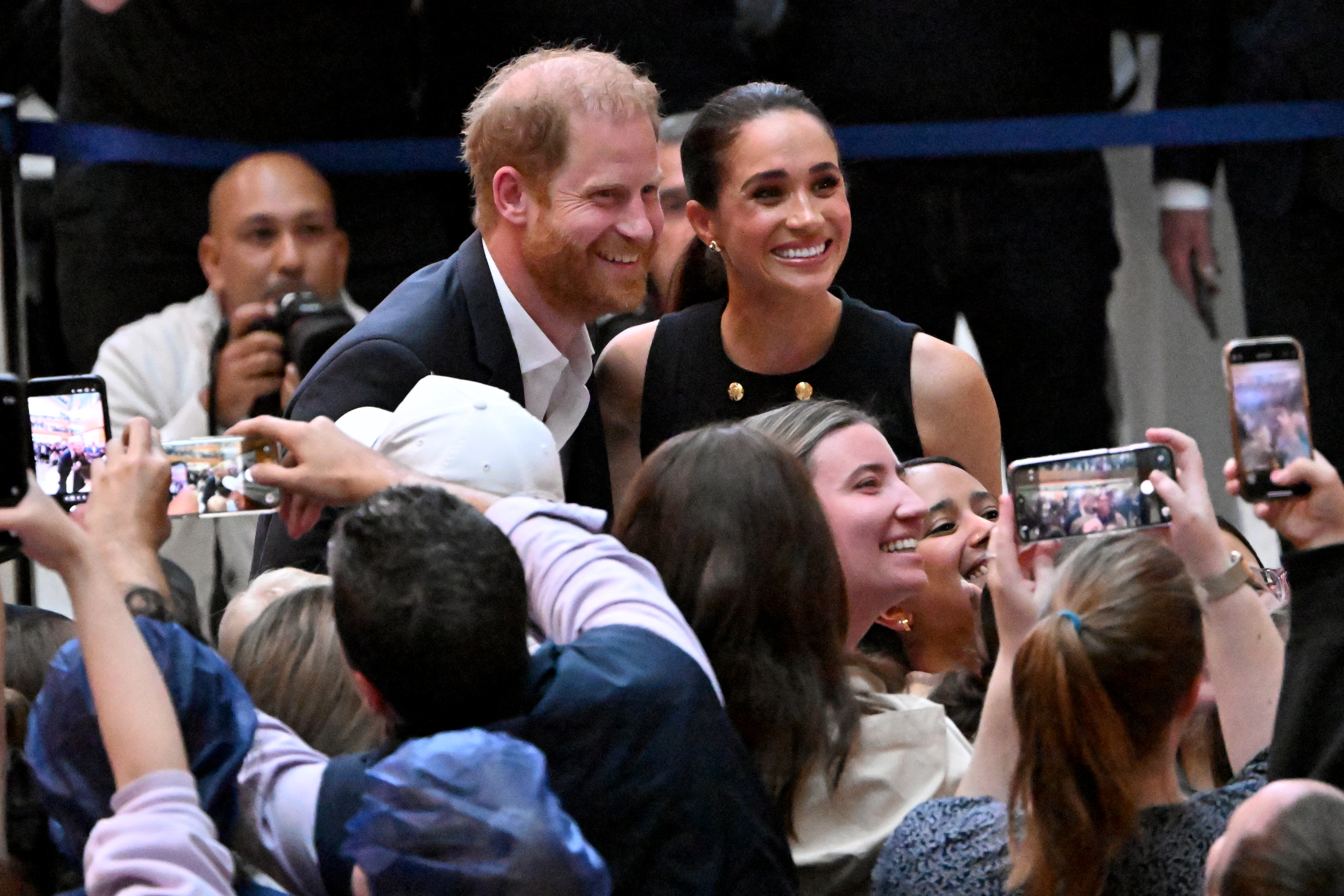 Prince Harry wears a baseball cap and a blue shirt, while Meghan Markle wears navy and white striped shirt and smiles