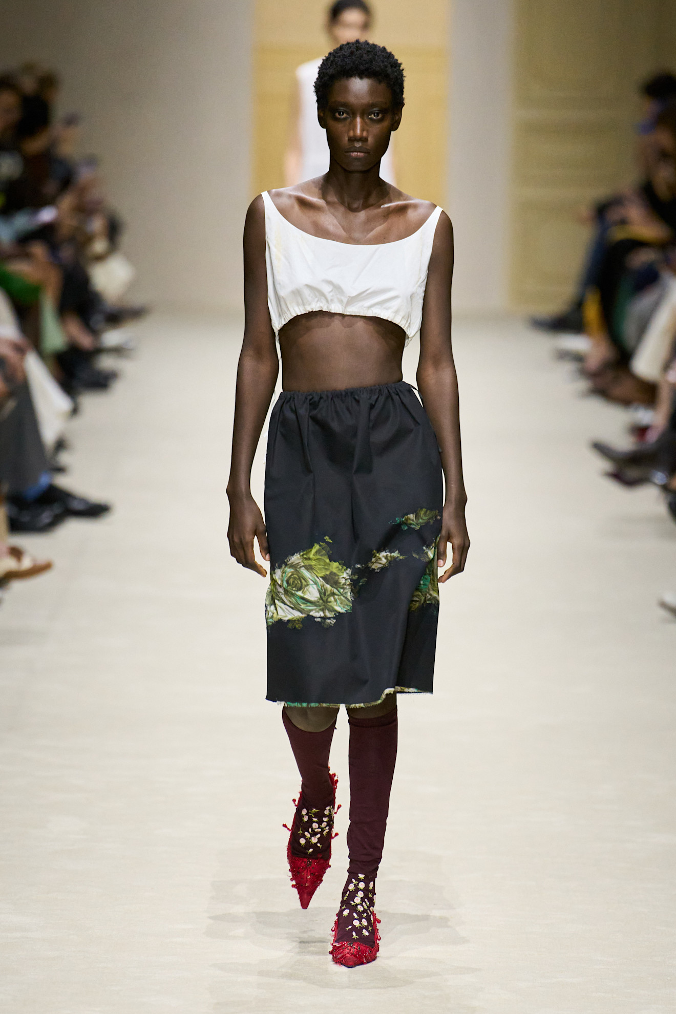 A model at the F/W 26 Prada show wearing a slasher skirt with a white crop top, knee-high socks, and red pumps.