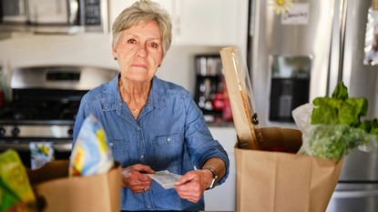 A senior woman in her kitchen with grocery bags, concerned about high prices.
