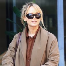 Jennifer lawrence wears black sunglasses, a tan coat, brown button down shirt, cognac leather bag, black pants, and black flats.