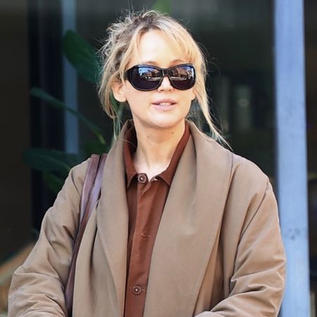 Jennifer lawrence wears black sunglasses, a tan coat, brown button down shirt, cognac leather bag, black pants, and black flats.