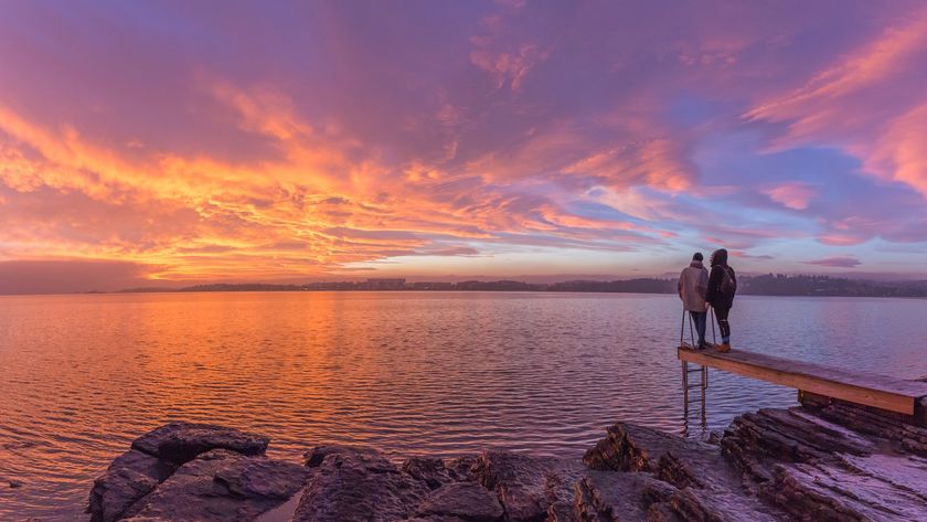 two people stand at the edge of a large body of water and look at the dramatic colorful sunset with the sky painted in golden light.