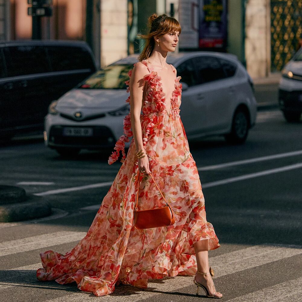 Woman wealks on a cross walk wearing a flowy flora dress that looks formal while carrying a handbag. 