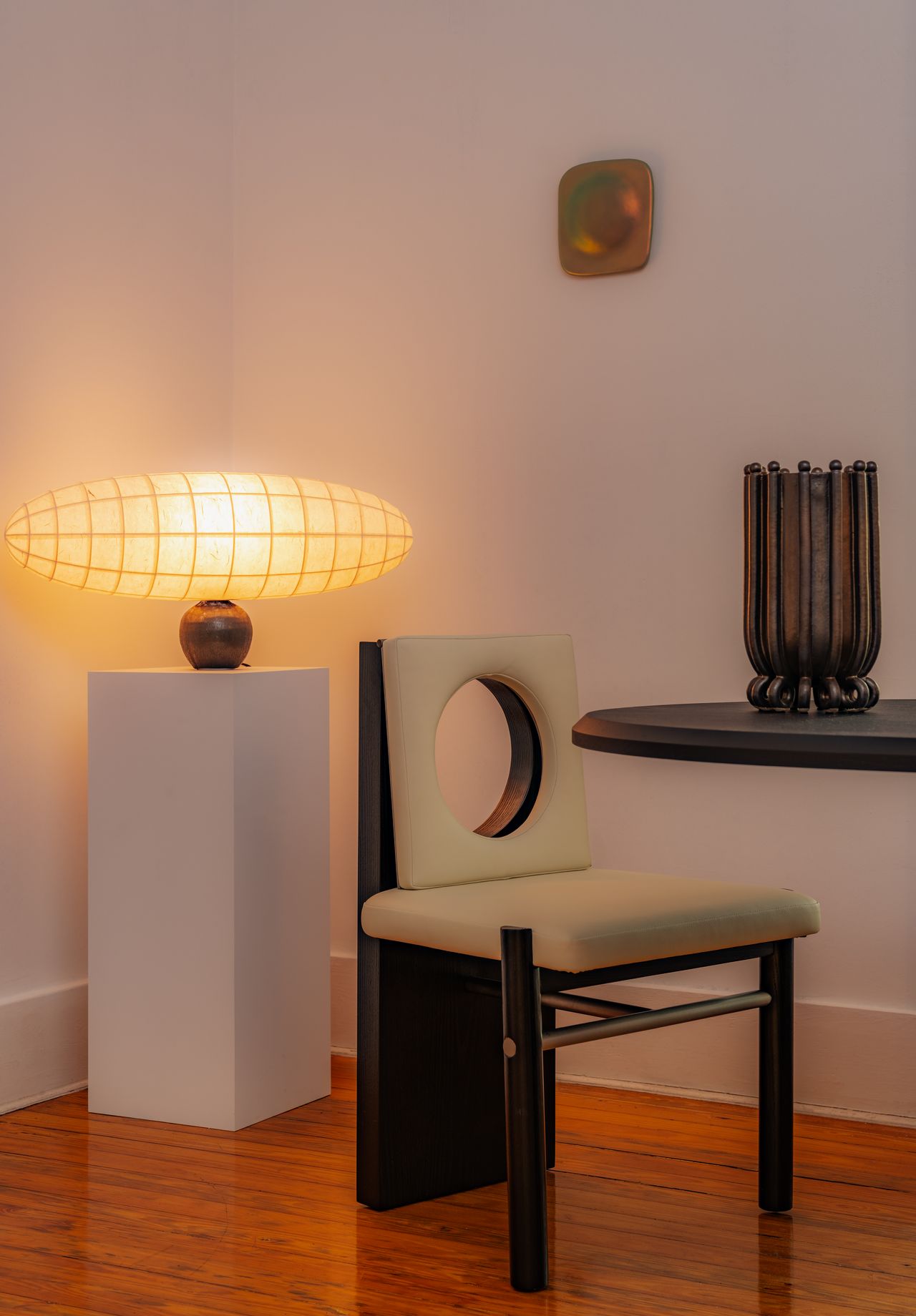 One oblong, warmly lit light sits on a white podium next to a champagne leather-upholstered chair with a holed back, a table, a vase, and a wall artwork.