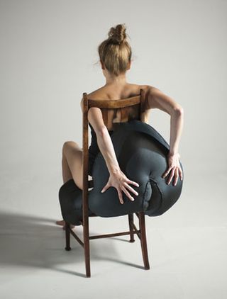 Naked female sitting on a wooden chair with a black padded section shaped like buttocks
