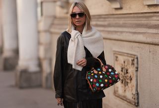 Karin Teigl is seen wearing double-framed sunglasses from Loewe; a cream-white knit sweater from Mango draped around her shoulders; a black leather jacket with long fringes combined with a black leather fringe midi skirt, both from Mango; a gold Oura ring combined with three different diamond rings; and a rectangular brown leather bag with logo pattern and multicolor painted dots from Louis Vuitton x Yayoi Kusama.