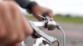 A close up of a silver Spurcycle Original bell, mounted to a silver handlebar, on which a white person's hands resting and slightly blurred
