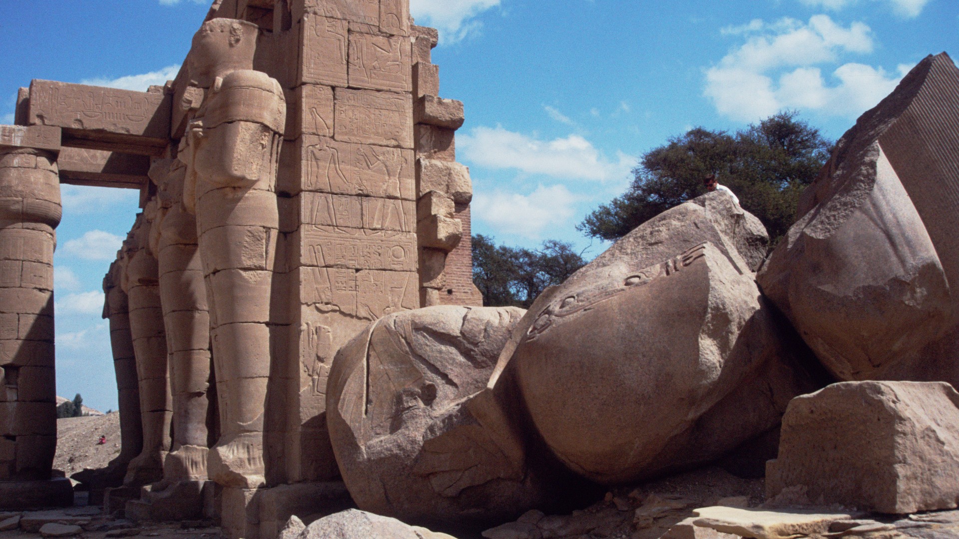 Ruínas de um antigo templo egípcio iluminado pelo sol, com imponentes colunas esculpidas e estátuas humanas. Na vanguarda está uma estátua gigante caída e quebrada em pedaços.