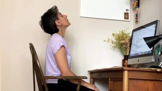 Fitness editor Yanar Alkayat is sat in a chair at her desk performing a neck stretch. Her hands rest in her lap and her head is tilted upwards, creating a stretch in her neck. In front of her is a Mac desktop computer and dried flowers in a case.