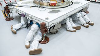 four pairs of feet covered in clean room booties protrude from underneath a spacecraft and its support stand
