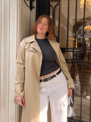 mid-size fashion influencer Raeann Langas leans outside a building door wearing a trench coat, cropped black tee, statement black belt, mini silver bag, and white cargo pants