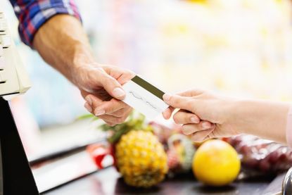 A man's hand holds a rewards credit card to get cash back while at the grocery stores check out, and a clerk's hand reaches out to swipe the card.