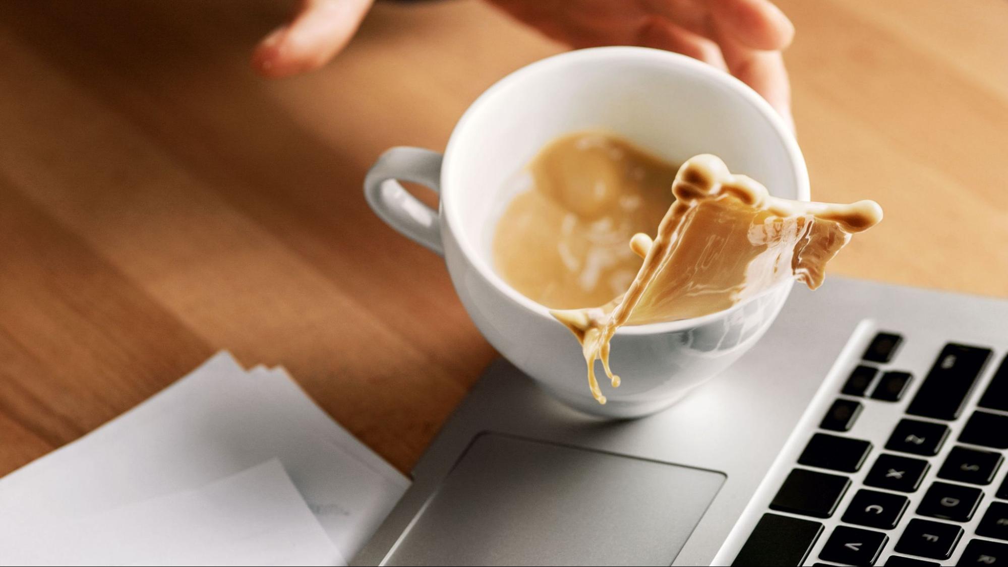 A hand drops a cup of coffee just above a laptop, with the coffee splashing over the side of the mug.
