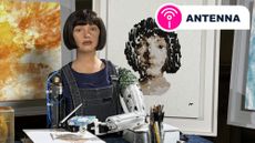 The Ai-Da robot sitting with a paintbrush in front of two paintings