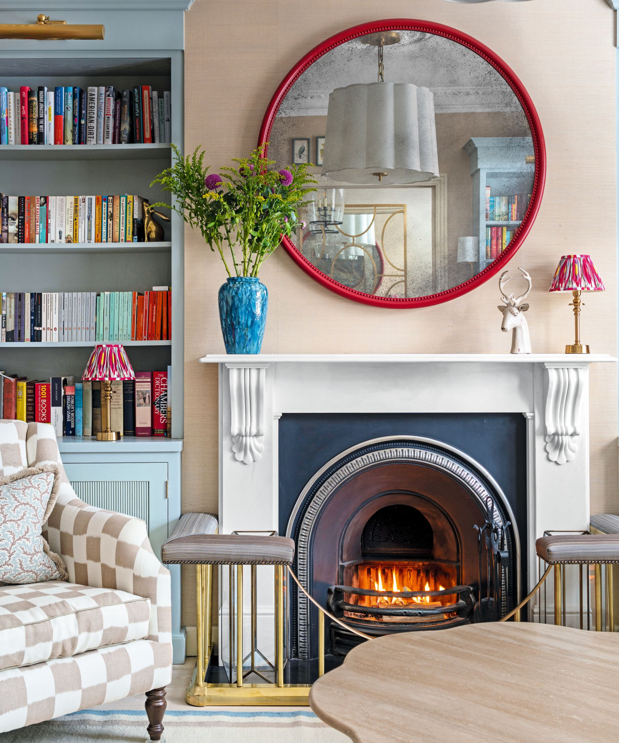 a small living room with a fireplace, a red mirror hanging above, a vase of flowers on the mantel, a bookshelf to the left, and a checker printed accent chair
