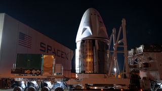 a white cone-shaped capsule on a truck bed in front of a large hangar with a logo reading &quot;spacex&quot; on it