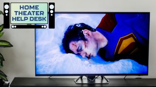 A TV on a credenza displaying a tattered, bloodied Superman from the 2025 movie "Superman." In the top-left corner of the image is a badge that reads: "Home Theater Help Desk."