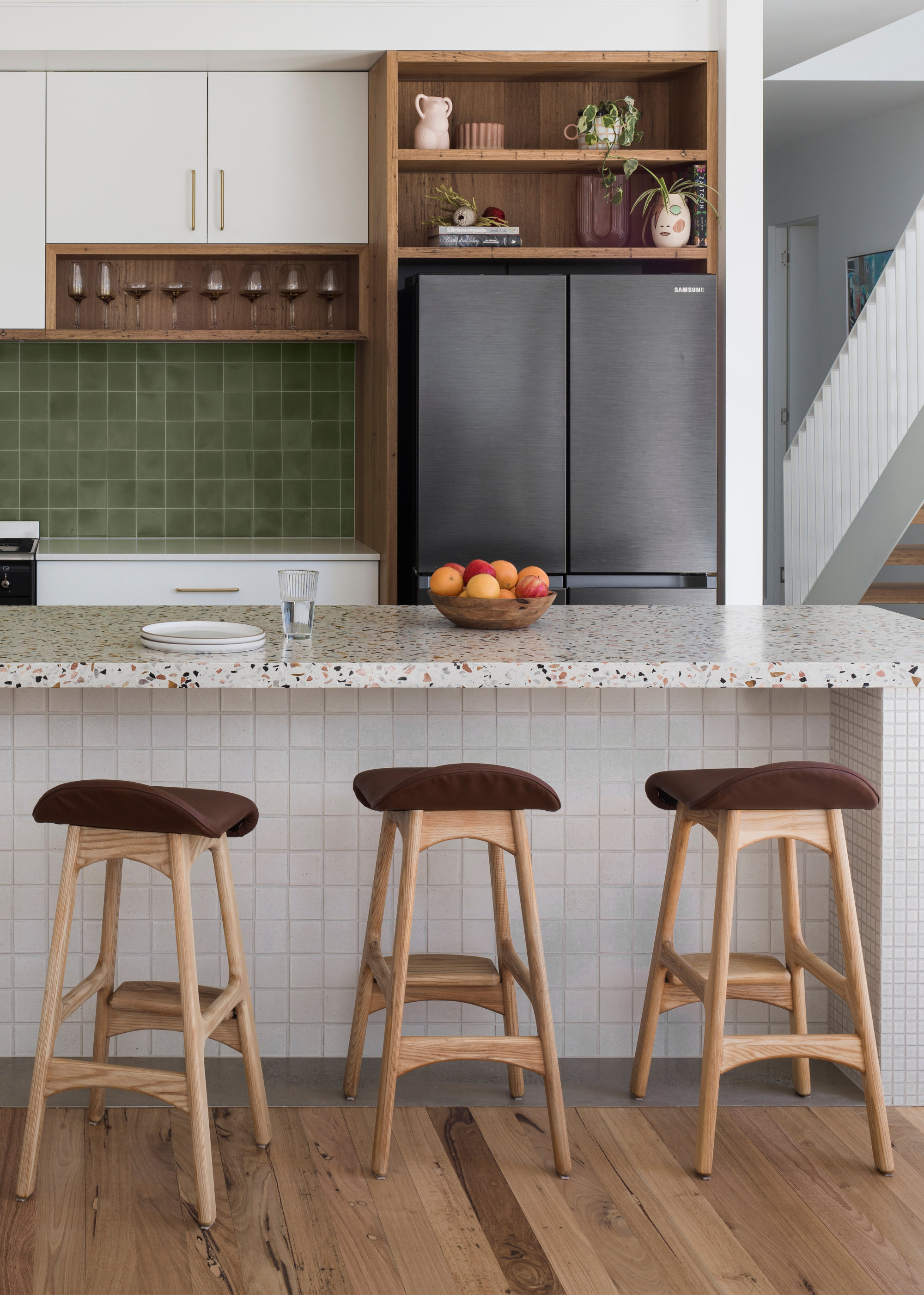 A kitchen with a green tiled backsplash a set of three stools by a tiled and terrazzo island and a fridge
