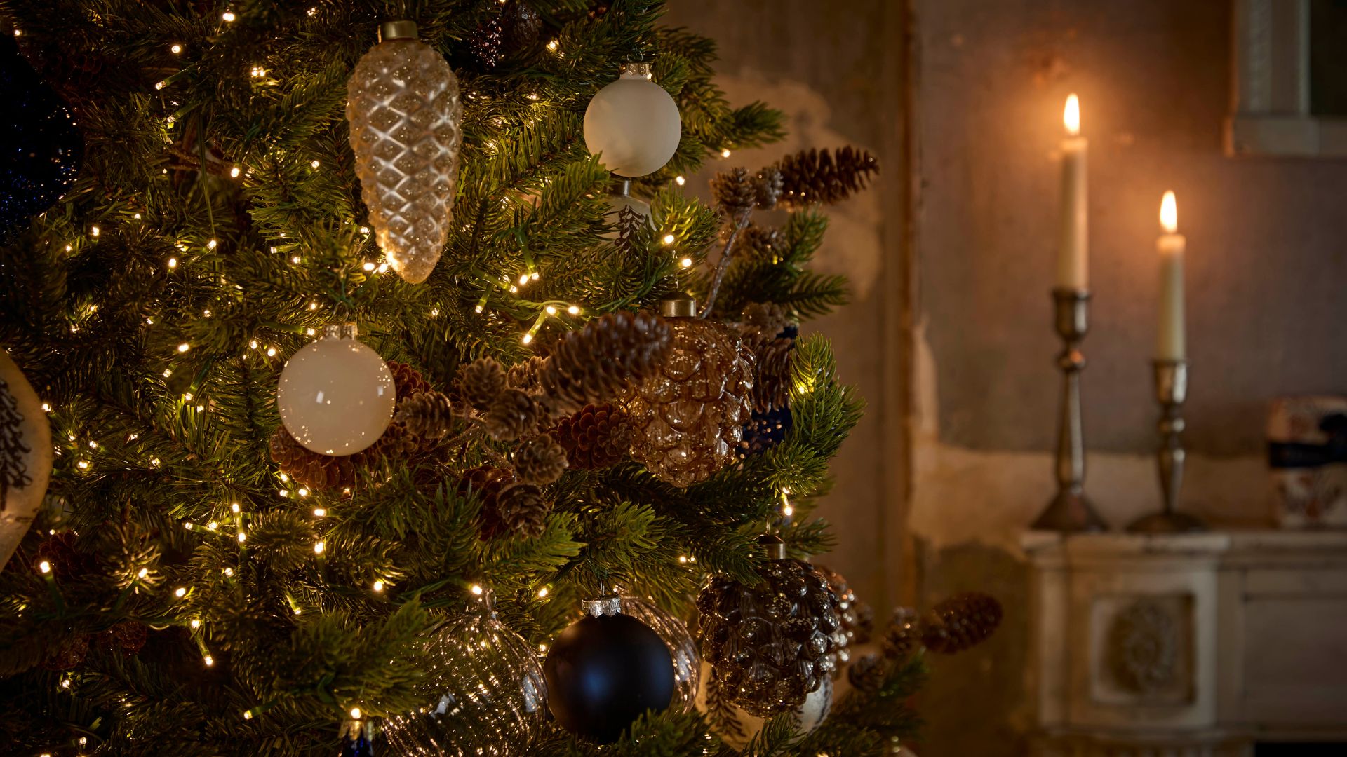 A close-up of a decorated Christmas tree by a mantel with lit tapers on holders
