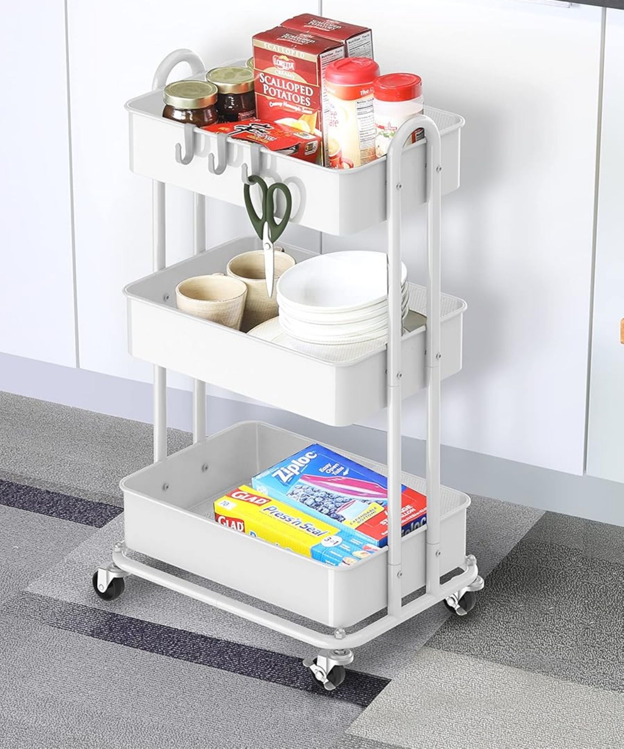 White utility rolling cart pictured in a white kitchen with grey flooring, filled with food, bowls, and other items.