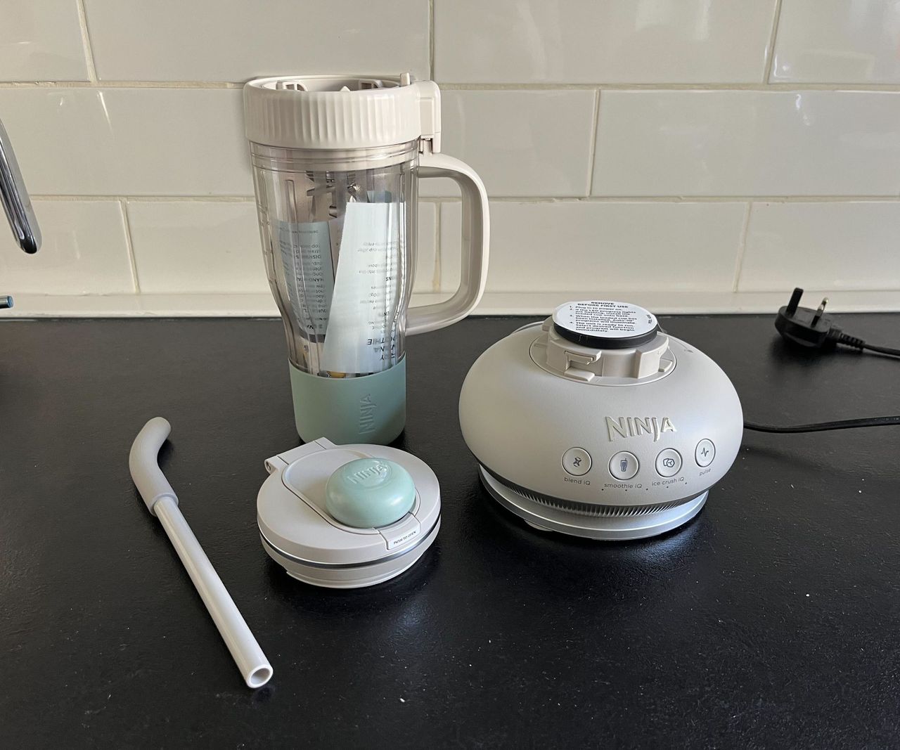 All the parts of a grey Ninja Blendboss, including the domed base, lid, staw, ad tumbler cup on a black counter.