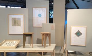 Geometric prints are framed and hung on a wall, with two wooden stools displayed on a platform in front of it.