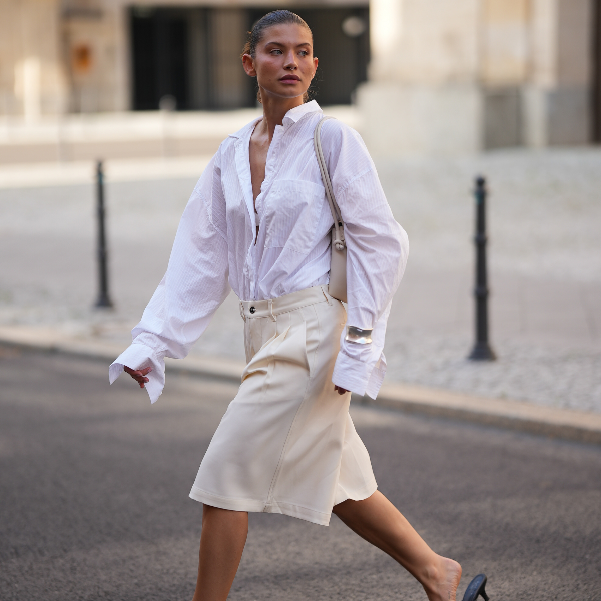 Sophia Geiss is seen wearing a white oversized shirt with subtle vertical stripes, a chest pocket and wide cuffs from Black Palms; cream white knee-length bermuda shorts with an oversized fit and front pleats from Eme Studios; black kitten heel thong sandals with a minimalist flip-flop silhouette and low heel from Leger by Lena Gercke; a soft ivory shoulder city bag with a folded flap, elongated shape and knotted strap details from comma Fashion; a silver chunky cuff bracelet worn above the shirt sleeve from COS; her brown hair is styled in a sleek low bun on July 27, 2025 in Berlin